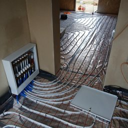 ART-INSTALBUD Artur Popowicz - Rozłożone rury ogrzewania podłogowego na folii izolacyjnej z widocznym rozdzielaczem w białej szafce i skrzynką przyłączeniową, w surowym wnętrzu budynku.