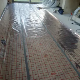 Mata grzewcza z siatką w czerwono-białą kratę, częściowo przykryta folią, przygotowana do montażu ogrzewania podłogowego w nowym budynku z dużym oknem tarasowym.