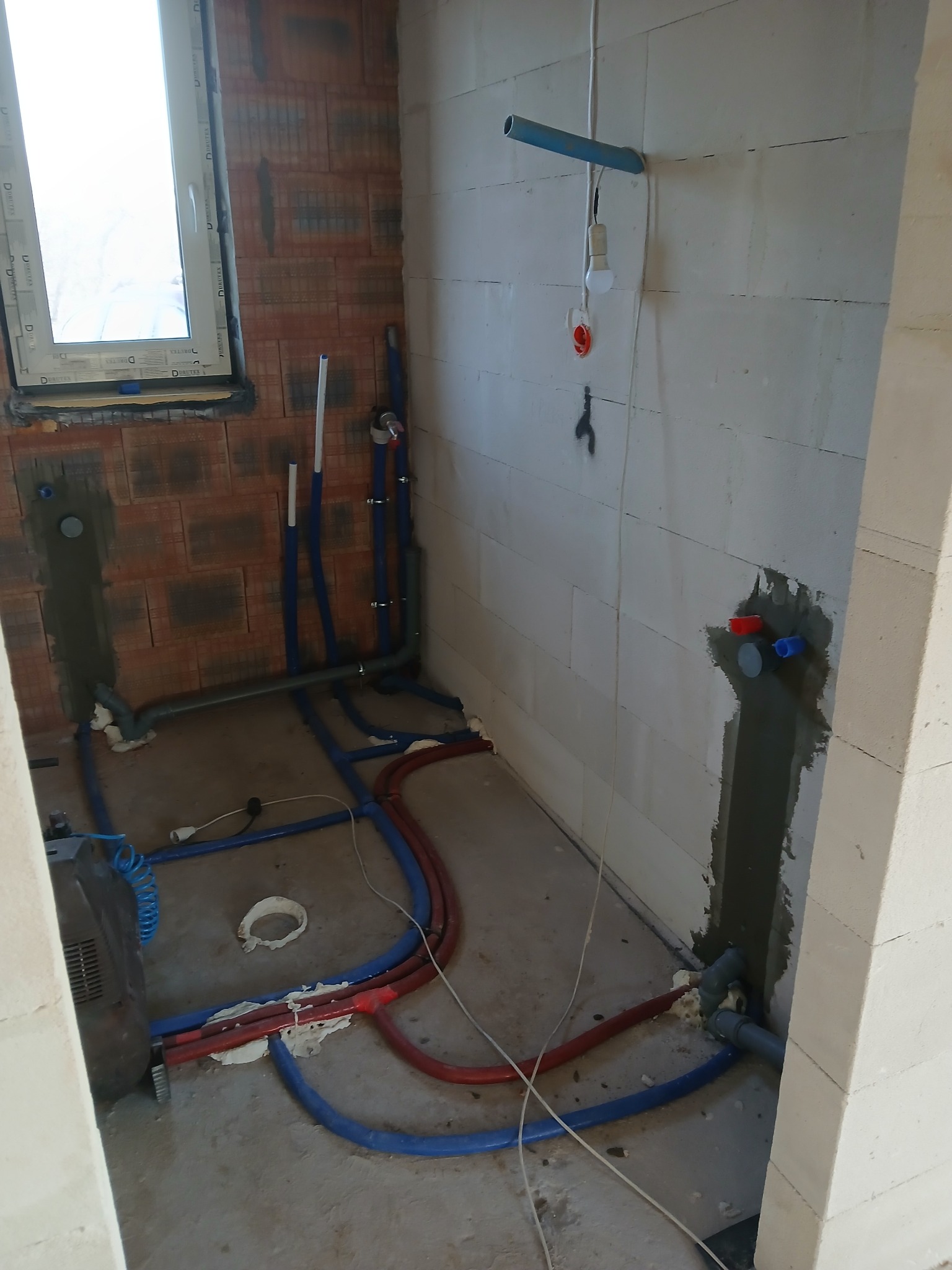 Surowy stan łazienki w trakcie prac hydraulicznych: widoczne rury instalacji wodnej i grzewczej w kolorze niebieskim i czerwonym, wystające ze ścian z betonu komórkowego i cegły, podłączone...