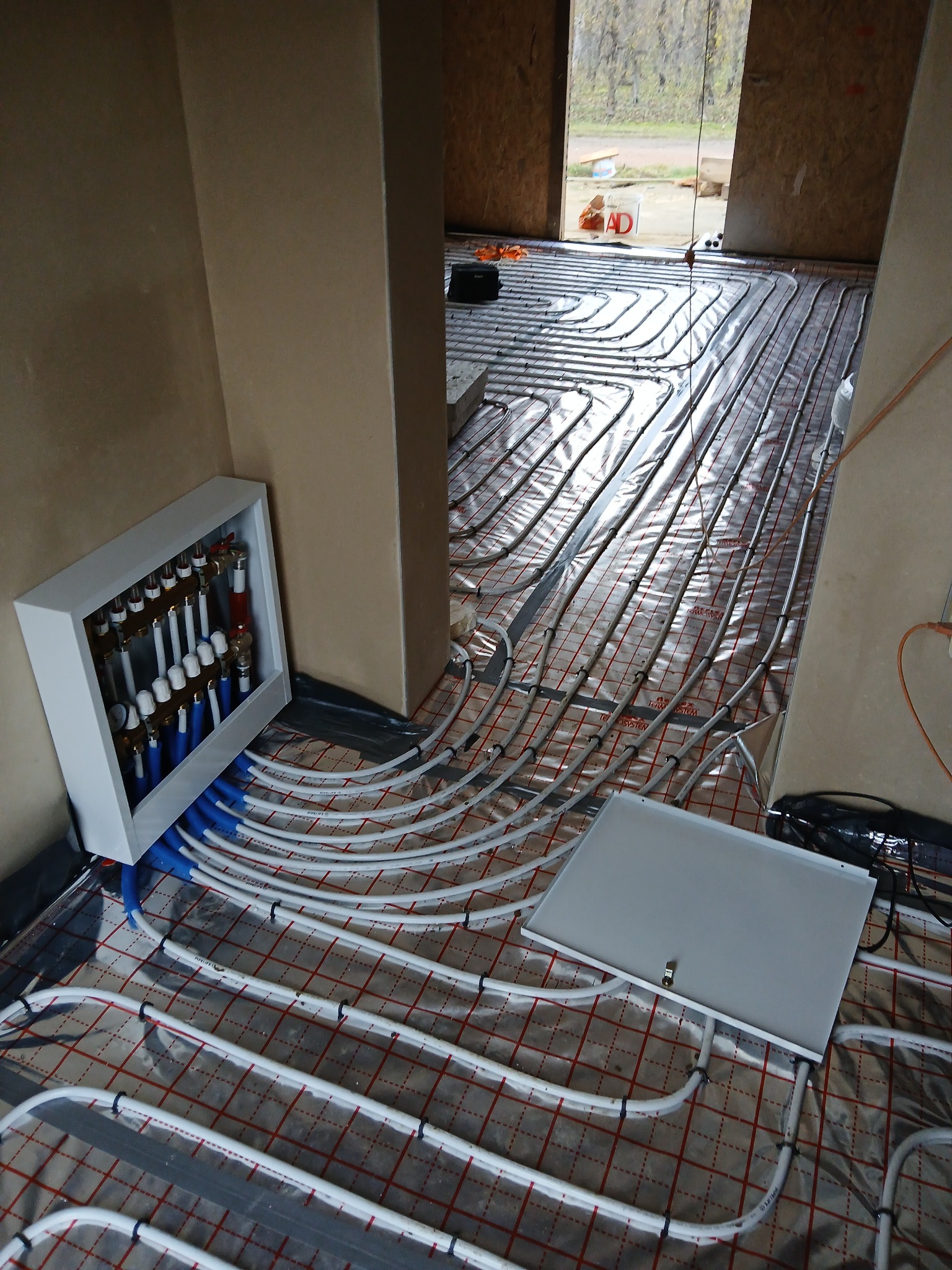 Rozłożone rury ogrzewania podłogowego na folii izolacyjnej z widocznym rozdzielaczem w białej szafce i skrzynką przyłączeniową, w surowym wnętrzu budynku.