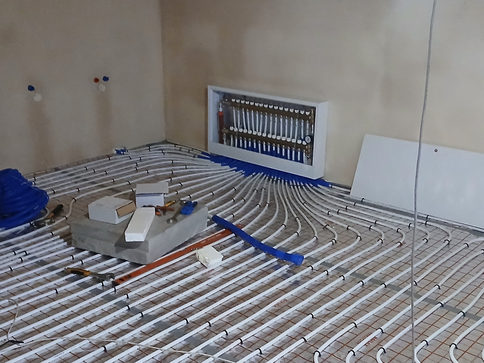 Rozłożone białe rurki ogrzewania podłogowego na siatce, z niebieskimi elementami mocującymi, widoczny rozdzielacz w białej obudowie, narzędzia pracy i materiały budowlane na betonowym bloku.