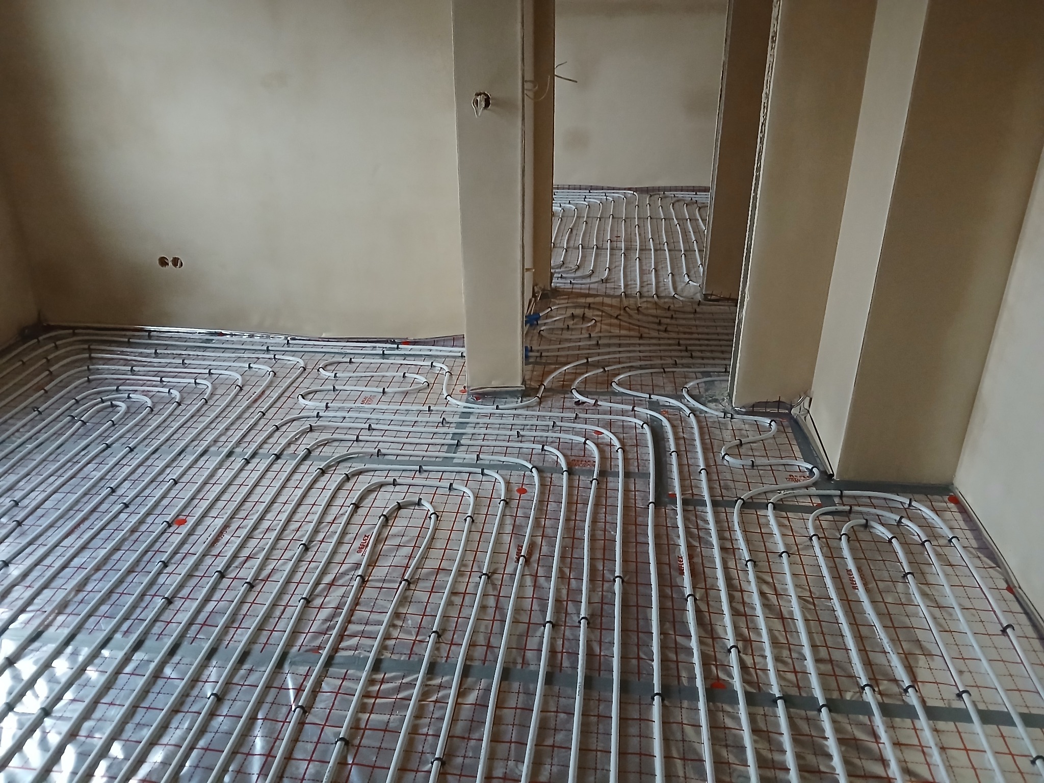 Białe rurki ogrzewania podłogowego rozłożone na folii z czerwoną siatką, widoczne w surowym stanie wnętrza z beżowymi ścianami i otworami na gniazdka elektryczne.