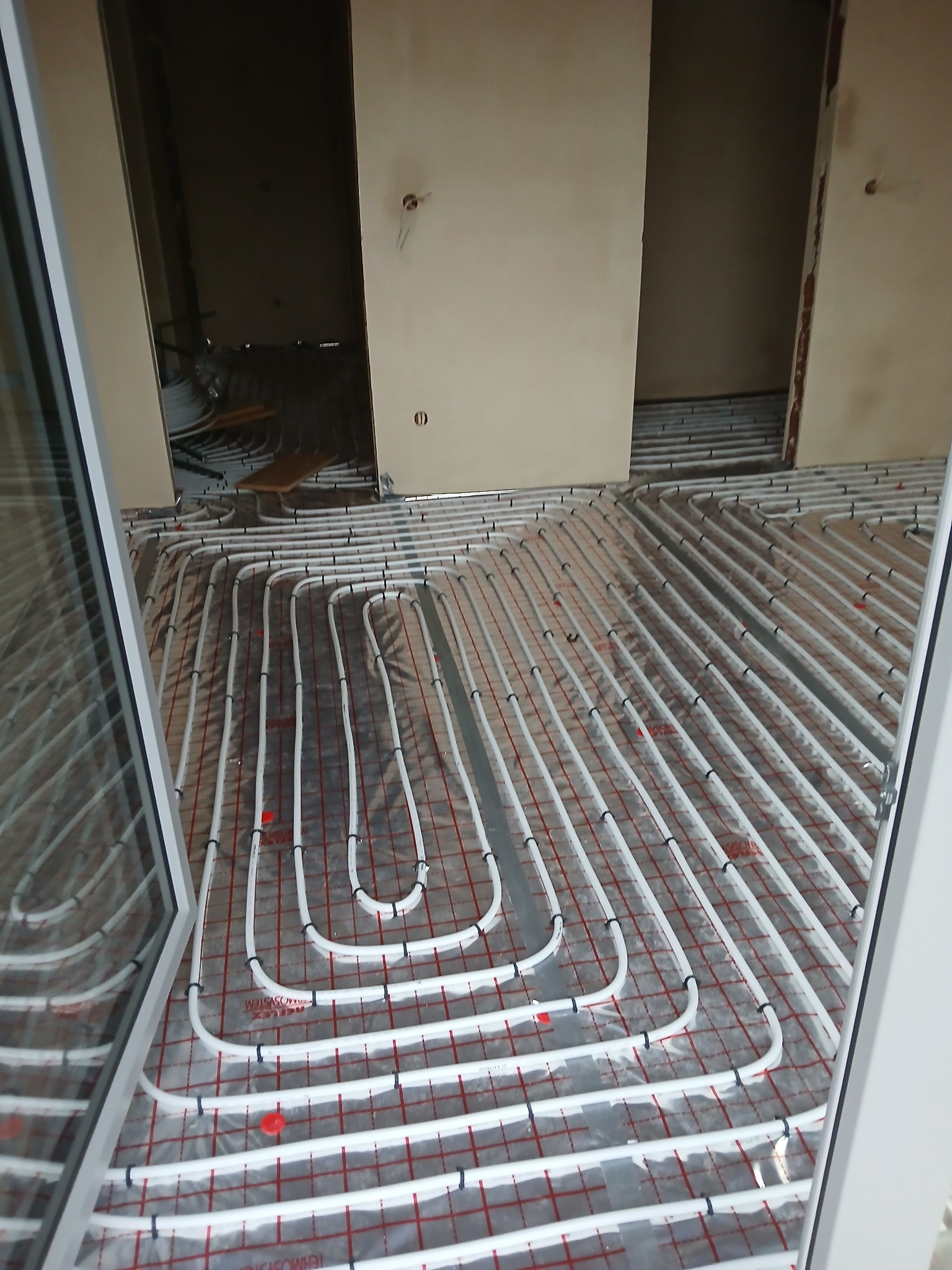 Białe rury ogrzewania podłogowego ułożone w pętle na folii z czerwoną siatką, widoczne przez otwarte okno, w surowym wnętrzu budynku.