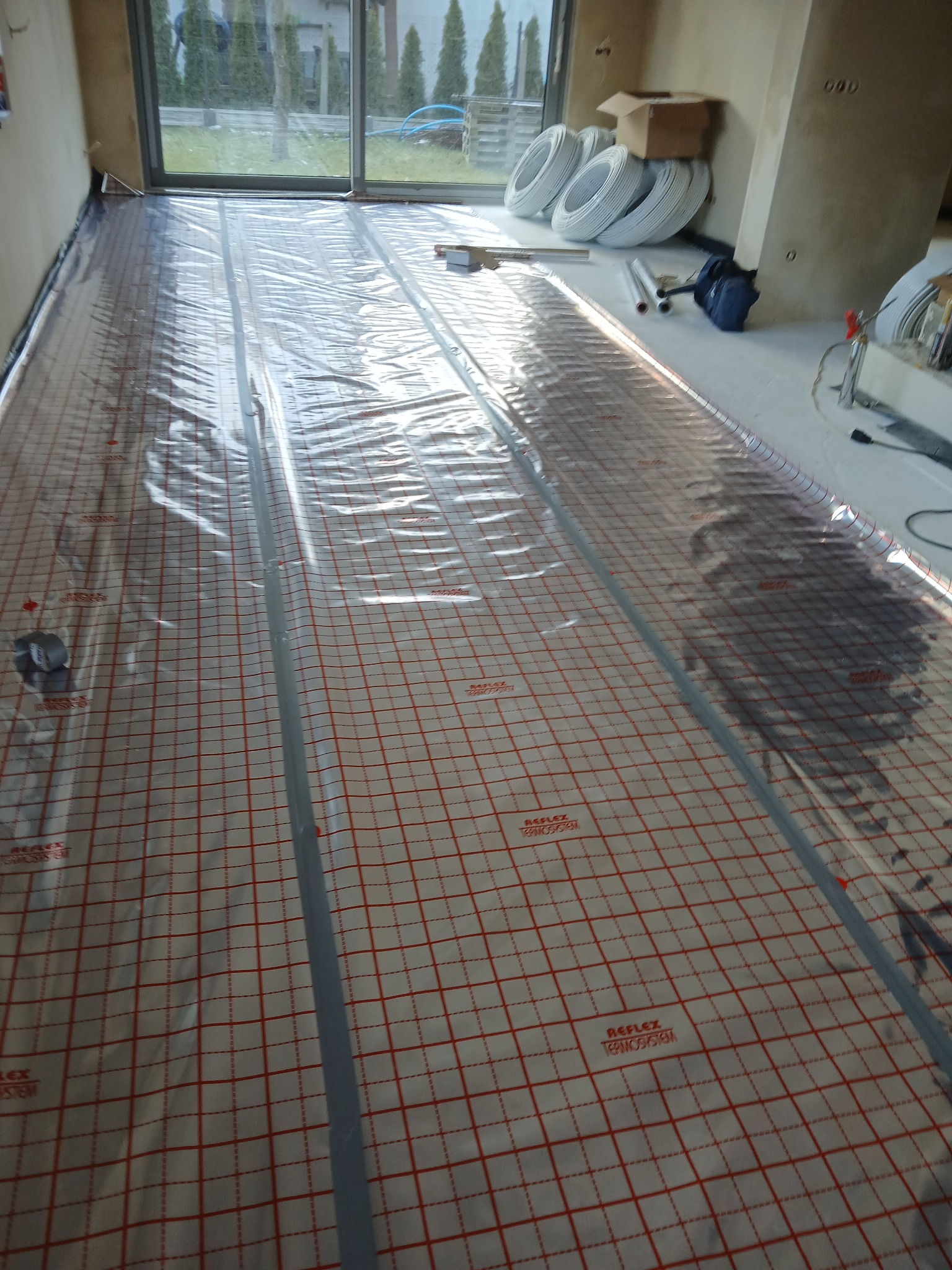 Mata grzewcza z siatką w czerwono-białą kratę, częściowo przykryta folią, przygotowana do montażu ogrzewania podłogowego w nowym budynku z dużym oknem tarasowym.