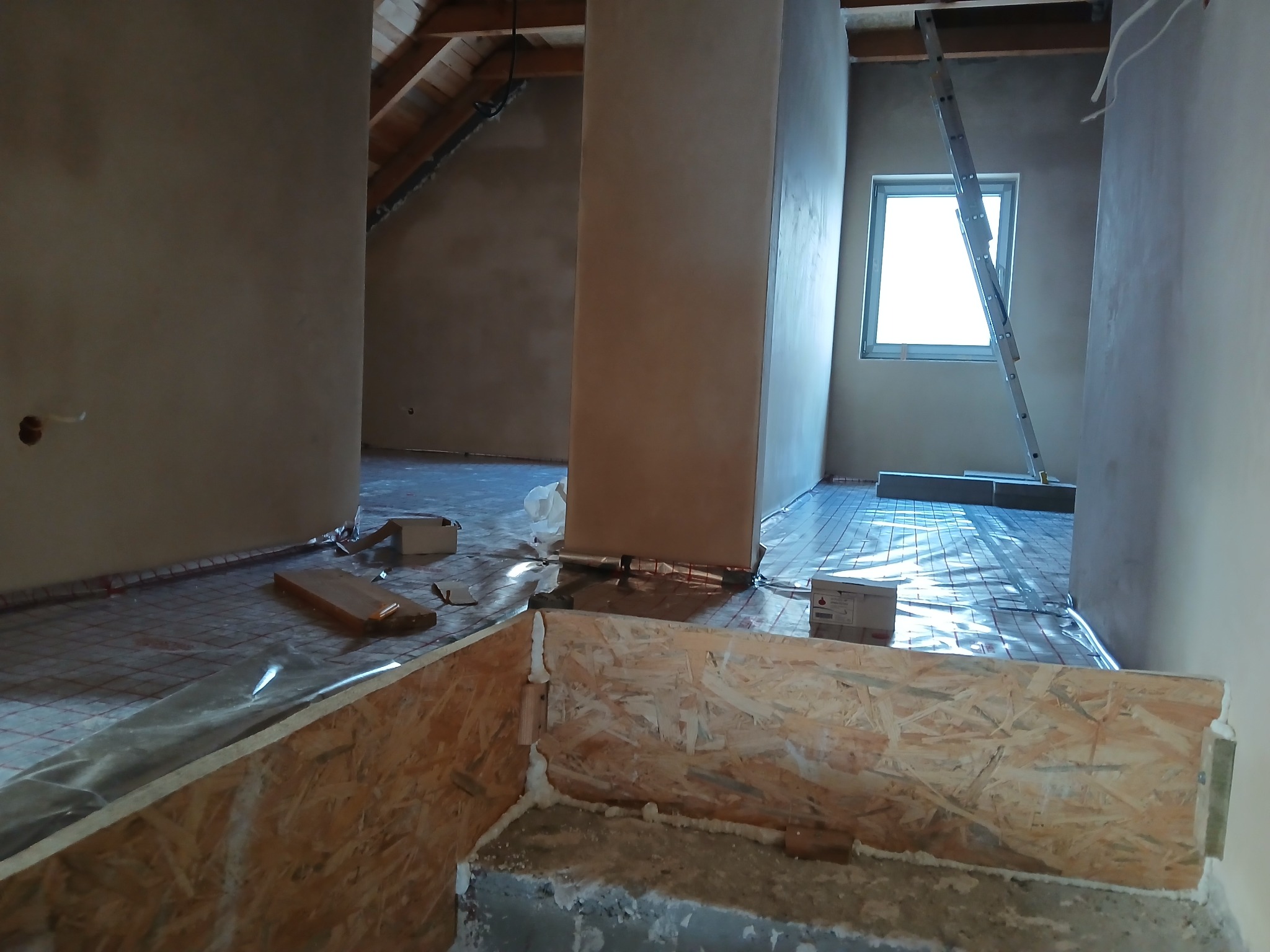 Wnętrze poddasza w trakcie montażu ogrzewania podłogowego, widoczne rury grzewcze rozłożone na podłodze i przykryte folią, oraz fragment konstrukcji z płyty OSB z pianką montażową.