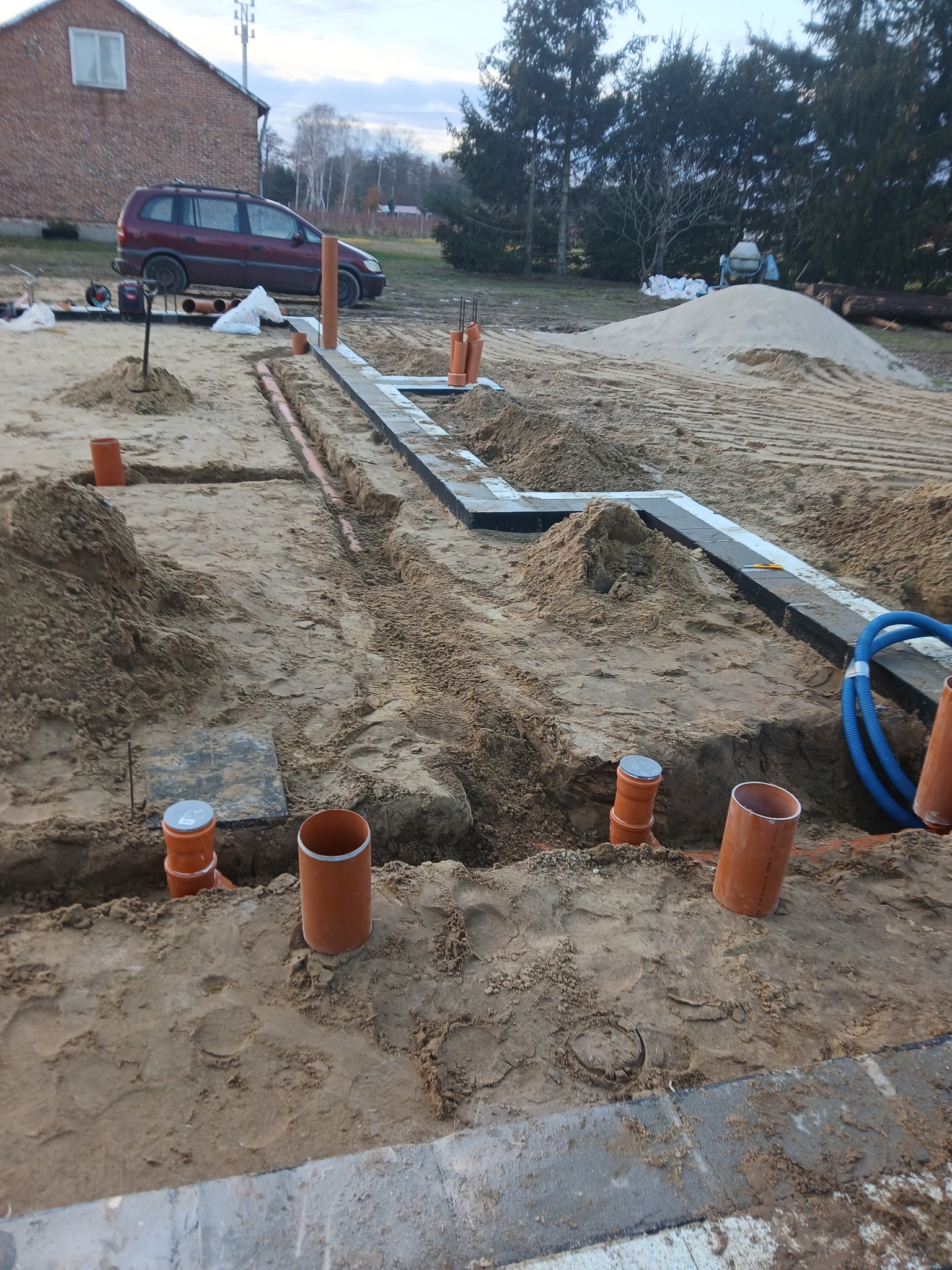 Wykop pod fundamenty z widocznymi rurami kanalizacyjnymi i drenażowymi, częściowo zasypany piaskiem, w tle samochód i betoniarka.