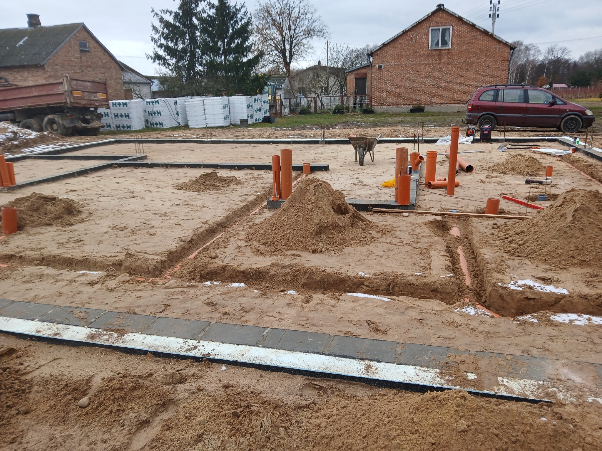Fundamenty domu z rozprowadzonymi rurami kanalizacyjnymi w wykopach, widoczny wózek budowlany i materiały budowlane H+H w tle.