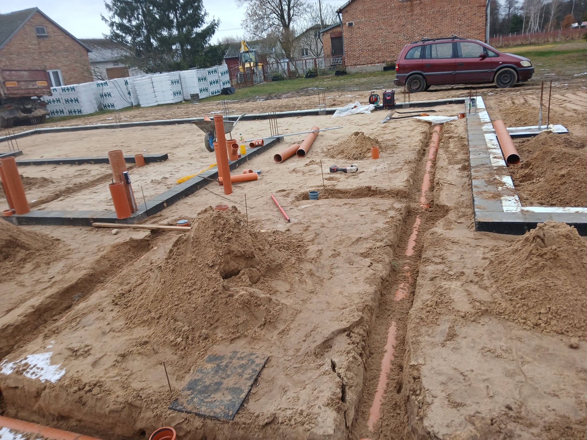Fundamenty domu w trakcie budowy z widocznymi rurami kanalizacyjnymi w wykopach, przygotowane do zasypania piaskiem, w tle ceglany budynek i zaparkowany samochód.