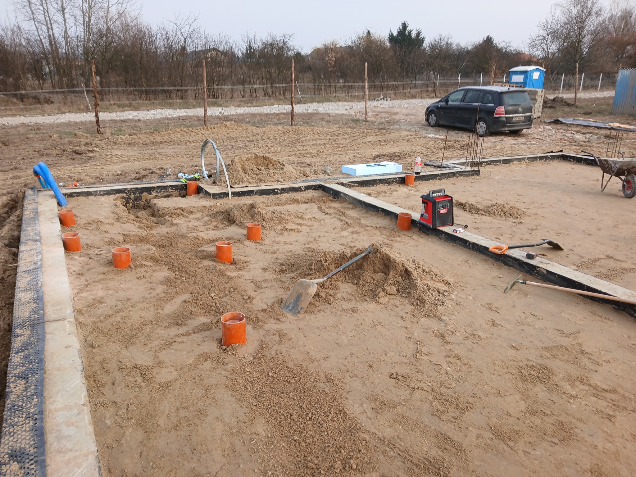 Przygotowanie terenu pod fundament z częściowo ułożonymi rurami kanalizacyjnymi i pomarańczowymi osłonami, widoczna łopata i inne narzędzia na placu budowy.