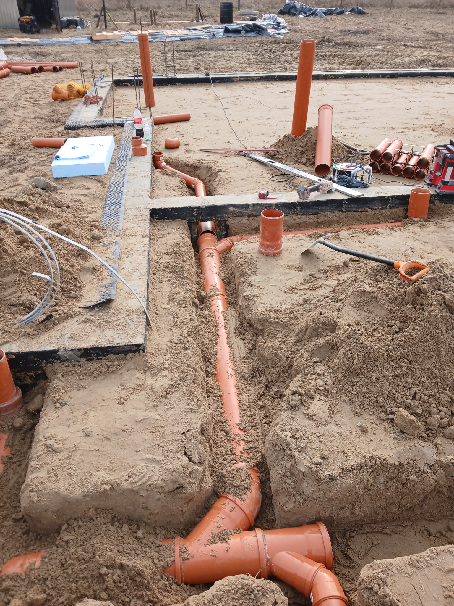 Pomarańczowe rury kanalizacyjne ułożone w wykopie, widoczne narzędzia i materiały budowlane na piaszczystym podłożu placu budowy.