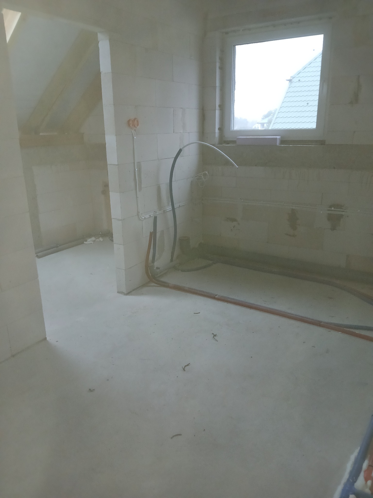 Surowy stan wnętrza z instalacją hydrauliczną w trakcie montażu, widoczne rury na ścianach i podłodze, okno z widokiem na dach sąsiedniego budynku.