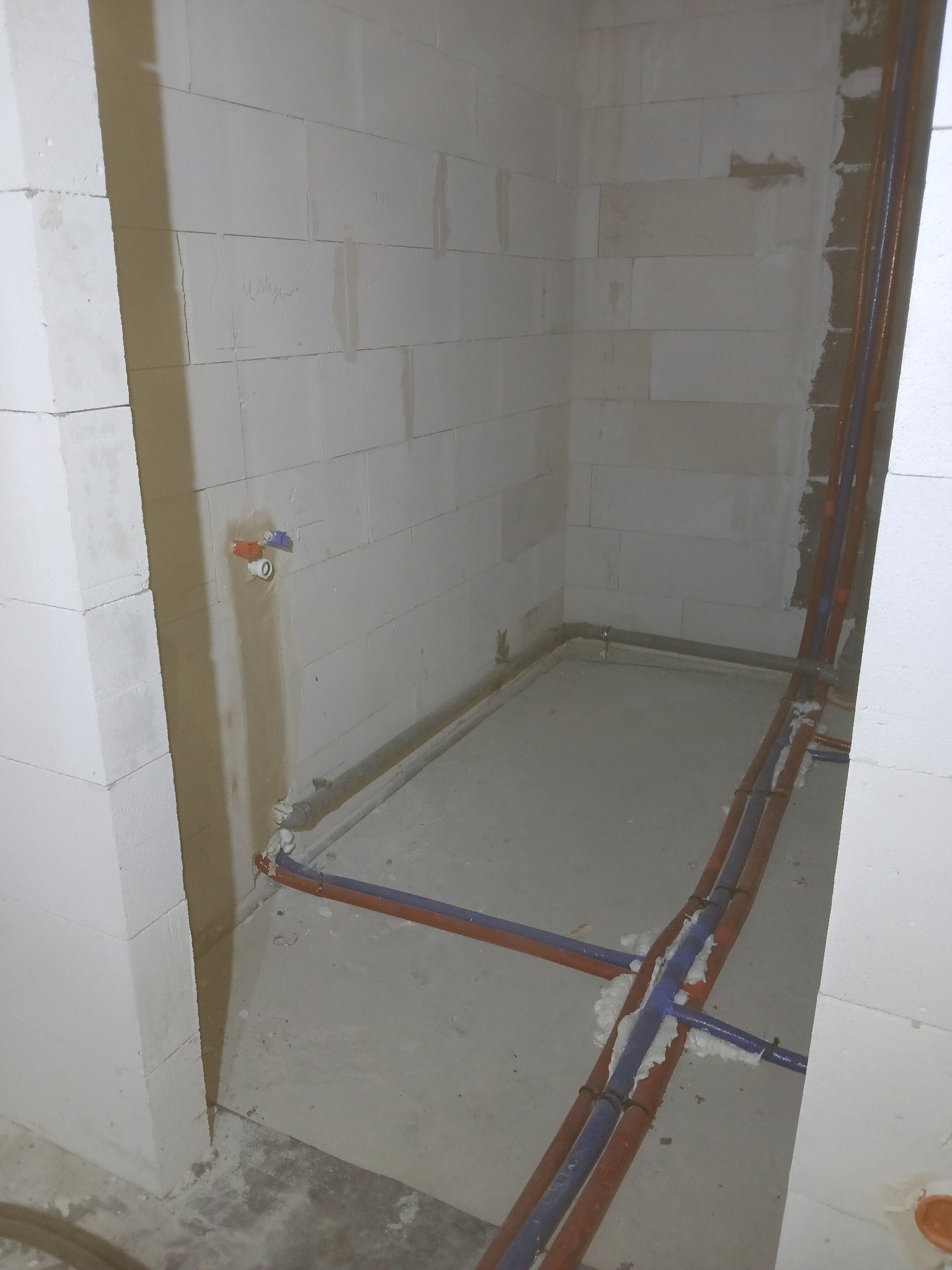 Surowy stan łazienki w trakcie prac instalacyjnych: widoczne rury wodne i kanalizacyjne prowadzone po ścianach z bloczków, zabezpieczone pianką montażową w miejscach łączeń, z przygotowanym...