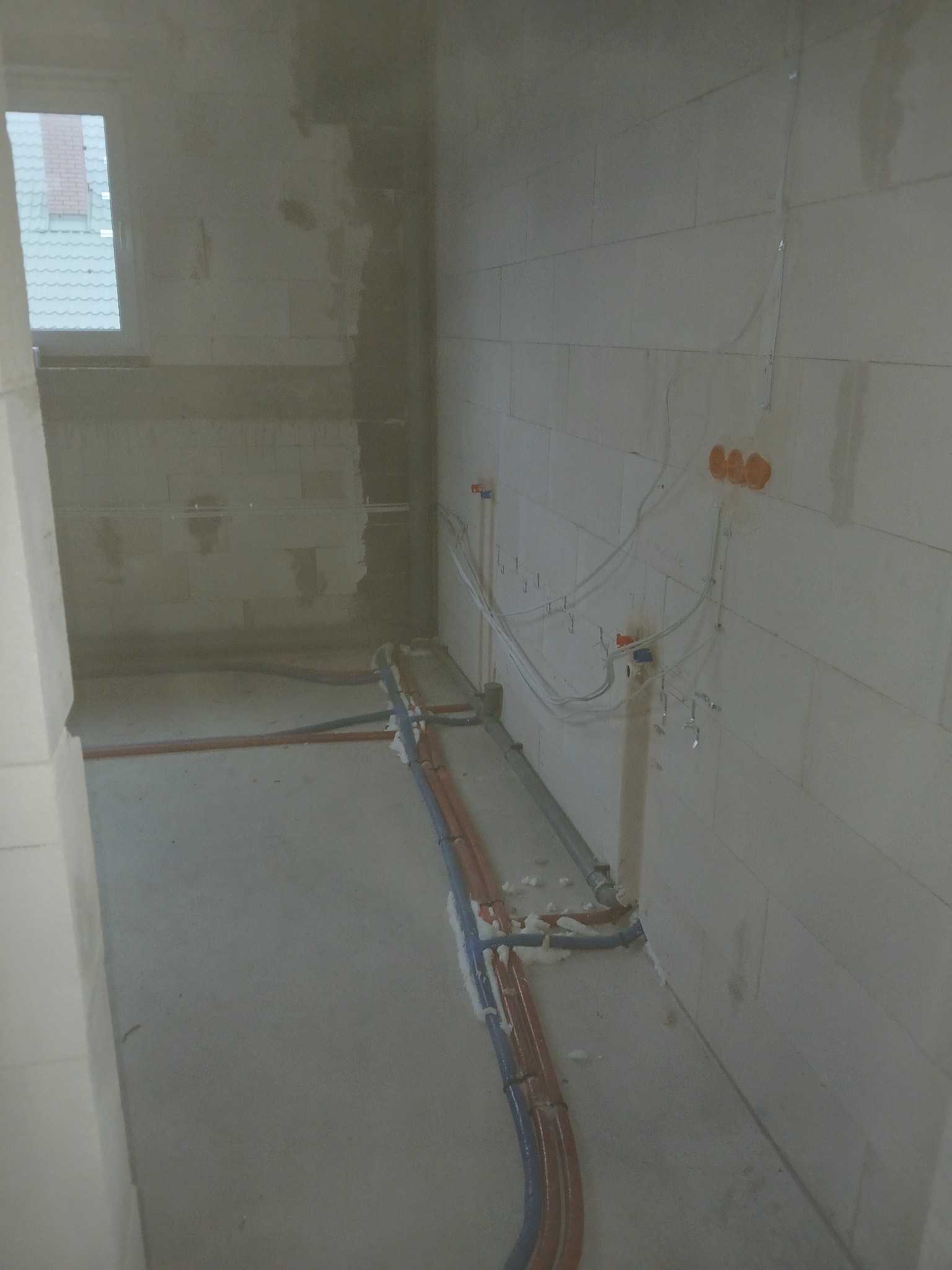 Wnętrze w stanie surowym z widocznymi rurami instalacji wodnej i grzewczej rozprowadzonymi po podłodze i ścianach, zabezpieczonymi pianką montażową.