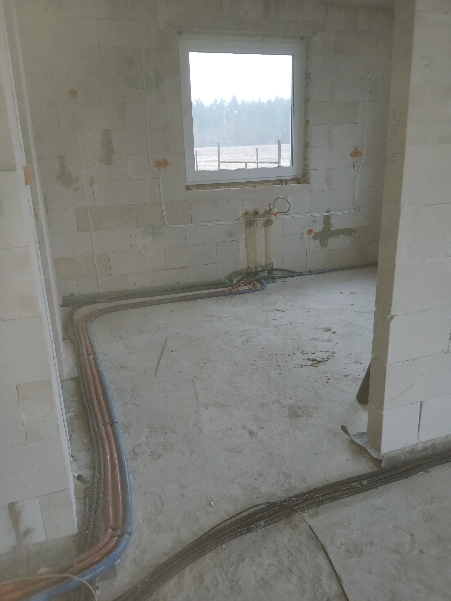 Surowy stan wnętrza z widocznymi rurami instalacji grzewczej i hydraulicznej w podłodze, oraz przewodami elektrycznymi w ścianach z betonu komórkowego w trakcie realizacji.