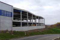 Surowy szkielet budynku w stanie deweloperskim, widok z boku, betonowe kolumny i belki, trawa na pierwszym planie, jesienne drzewa w tle, pochmurne niebo.