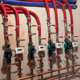 Lux Klima Oliwier Szymczak - Rozbudowana instalacja centralnego ogrzewania z rzędami miedzianych rur, pomp obiegowych Wilo i izolowanych rur doprowadzających w kolorach czerwonym i niebieskim, zamontowana na ścianie.