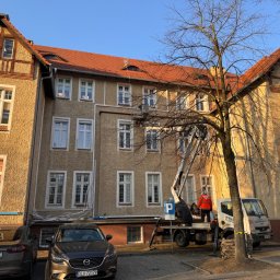 LUX KLIMA - Pracownik w pomarańczowej kurtce na podnośniku koszowym przy budynku z czerwonym dachem, prawdopodobnie montaż lub serwis instalacji na elewacji, widoczne kable i rury. Na pierwszym planie...