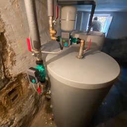 LUX KLIMA - Szary zbiornik buforowy z podłączoną armaturą, w tym zawory z czerwonymi uchwytami i zielony regulator, umieszczony w surowym pomieszczeniu z nierównymi ścianami.