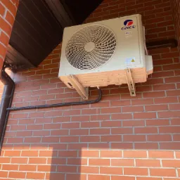 Zamontowana jednostka zewnętrzna klimatyzacji marki Gree na ceglanej ścianie budynku, z widocznymi elementami montażowymi i rurą odprowadzającą.