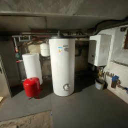 Lux Klima Oliwier Szymczak - Instalacja centralnego ogrzewania w piwnicy: białe zbiorniki akumulacyjne, czerwony zbiornik przeponowy, rury, zawory i osprzęt na tle surowych ścian i podłogi.