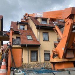 LUX KLIMA - Pomarańczowy podnośnik koszowy na tle domu z czerwoną dachówką, osoba w koszu podnośnika przy dachu, tabliczka ostrzegawcza na platformie podnośnika.