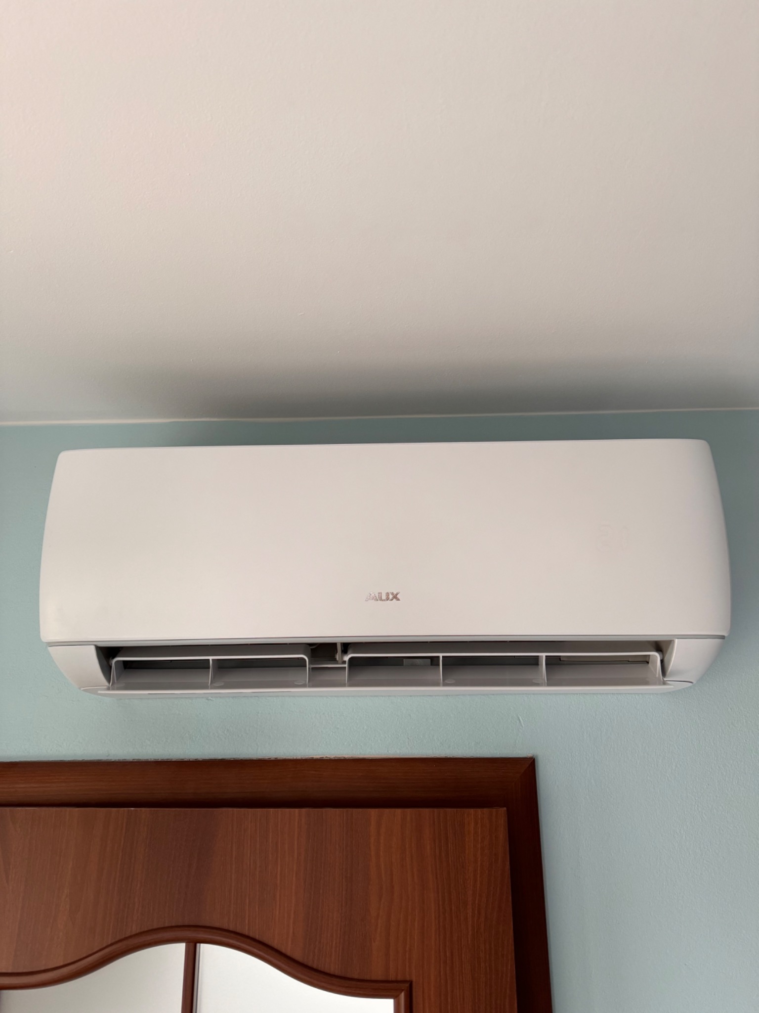 Biały klimatyzator AUX zamontowany na błękitnej ścianie nad brązowymi drzwiami.