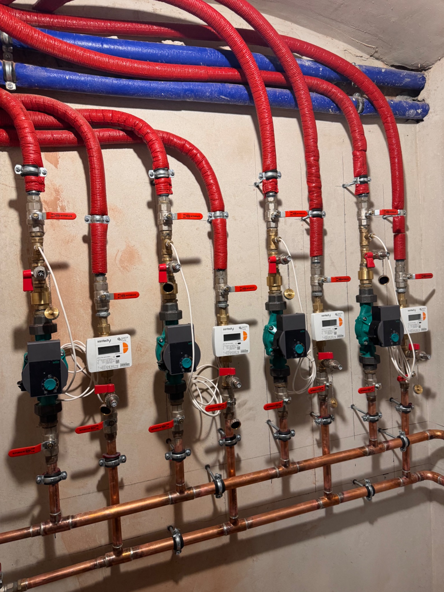 Rozbudowana instalacja centralnego ogrzewania z rzędami miedzianych rur, pomp obiegowych Wilo i izolowanych rur doprowadzających w kolorach czerwonym i niebieskim, zamontowana na ścianie.