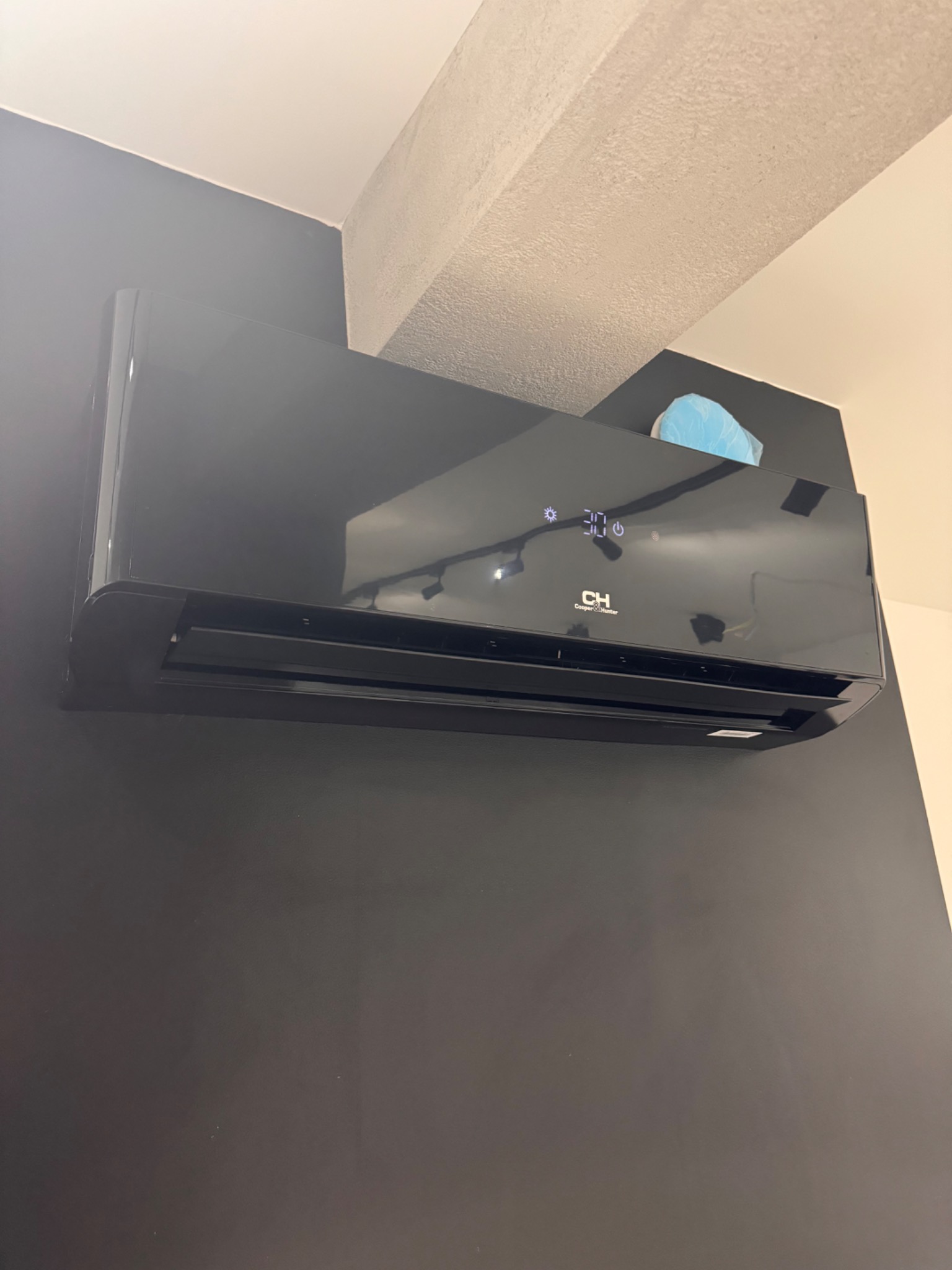 Czarna klimatyzacja ścienna Cooper&Hunter z wyświetlaczem ustawionym na 30 stopni, zamontowana na ciemnoszarej ścianie w rogu pomieszczenia z widoczną instalacją elektryczną w tle.