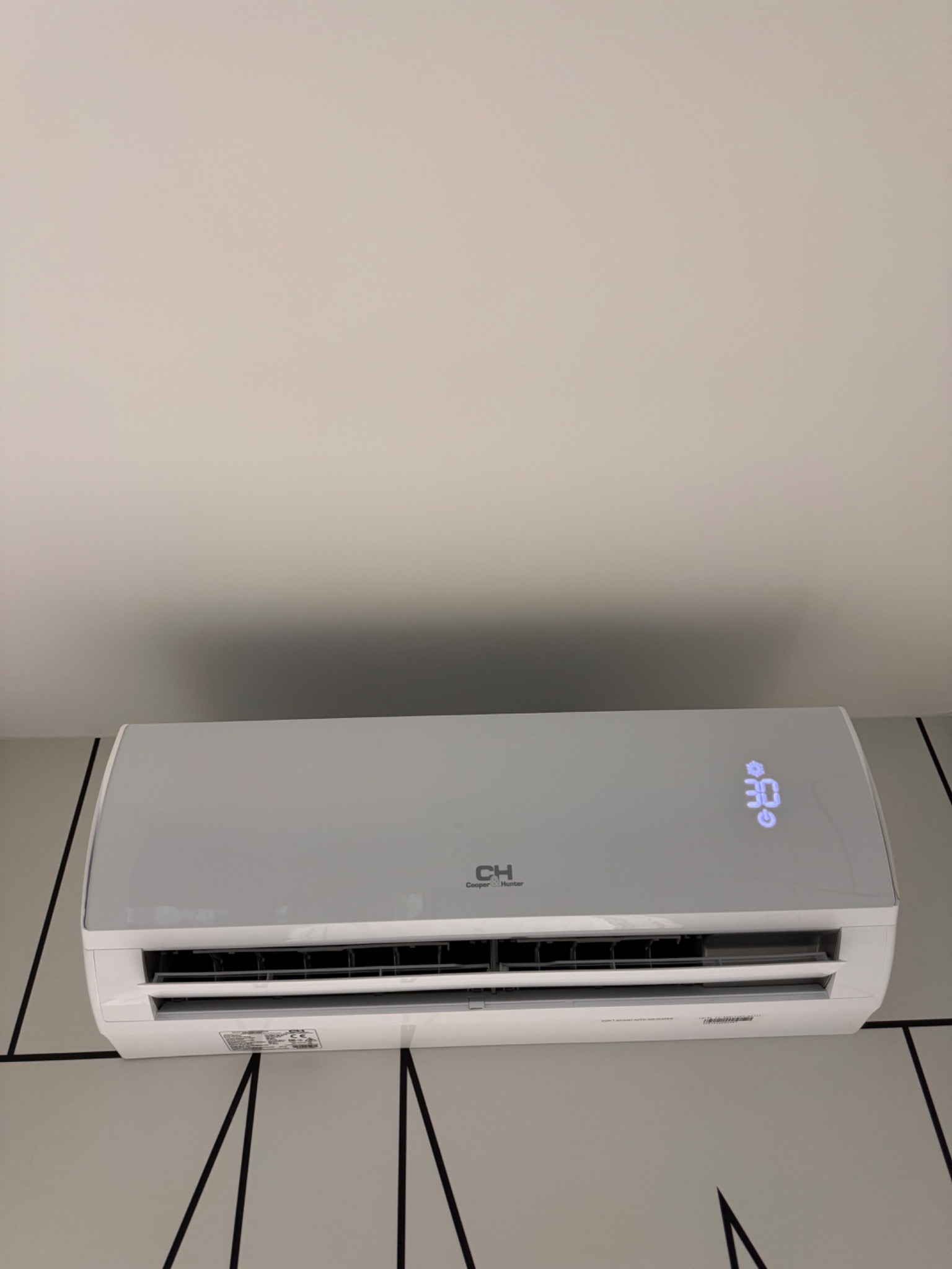 Biała klimatyzacja ścienna marki CH Cooper&Hunter z wyświetlaczem pokazującym temperaturę 30 stopni Celsjusza, zamontowana na ścianie z geometrycznym wzorem.