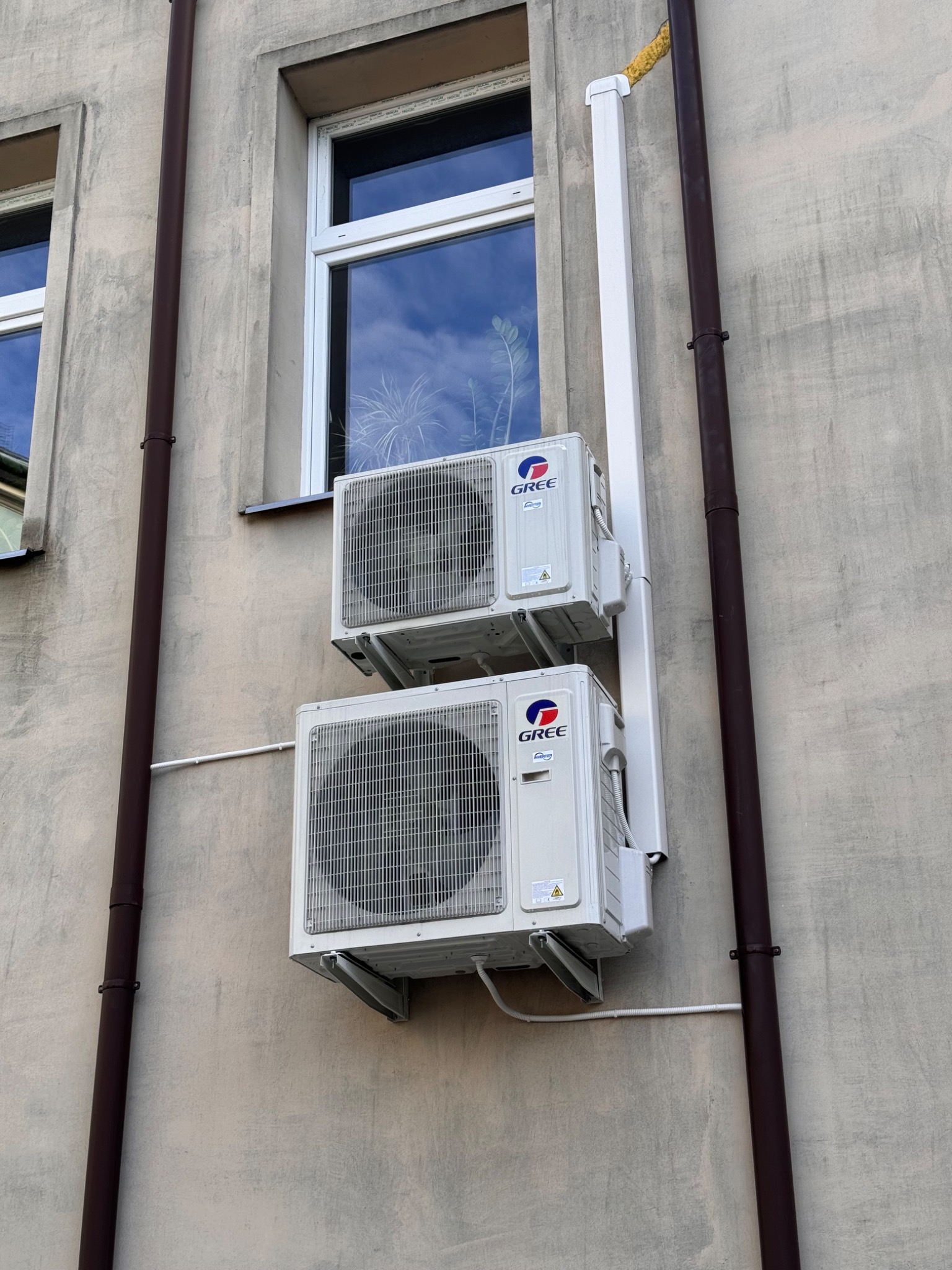 Dwa klimatyzatory marki Gree zamontowane jeden nad drugim na szarej ścianie budynku, obok brązowej rynny i białej rury odprowadzającej skropliny. W oknie odbija się zieleń.