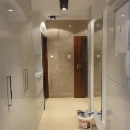 Nowoczesny przedpokój z białymi, lakierowanymi szafami w zabudowie, lustrami na ścianie, beżową ścianą z efektem marmuru oraz drewnianymi drzwiami. Wiadro z akcesoriami malarskimi stoi na podłodze.