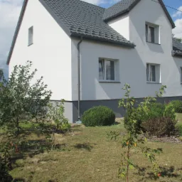 Dwukondygnacyjny dom jednorodzinny z białą elewacją i szarą podmurówką, widoczny z perspektywy ogrodu z młodymi drzewkami owocowymi, w tle taczka.