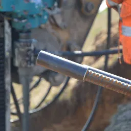 Wbijanie gwoździ gruntowych za pomocą specjalistycznej maszyny w województwie łódzkim, widoczny fragment pomarańczowego ubrania roboczego.