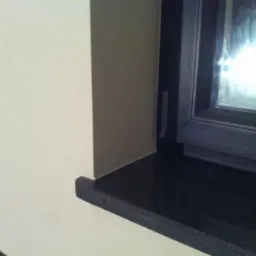 Gładka, beżowa ściana z narożnikiem i czarnym parapetem okiennym. Widoczny fragment okna. Wykończenie wnętrza w neutralnych barwach.