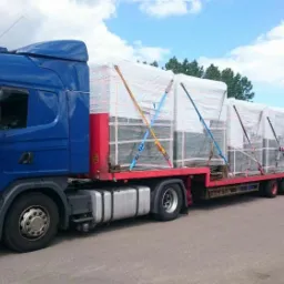 Niebieska ciężar&oacute;wka Scania z czerwoną platformą przewozi zabezpieczony ładunek na zewnątrz, w tle drzewa i budynki. Słoneczny dzień.