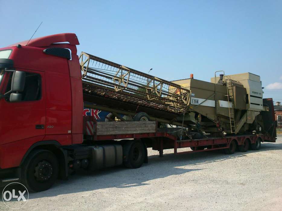 Czerwona ciężarówka przewozi kombajn zbożowy na niskopodwoziowej naczepie. Widoczne detale kombajnu i mocowanie ładunku.