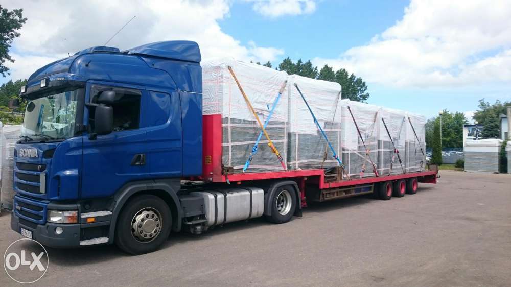 Niebieska ciężarówka Scania z czerwoną platformą przewozi zabezpieczony ładunek na zewnątrz, w tle drzewa i budynki. Słoneczny dzień.