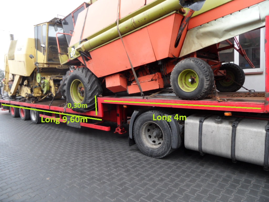 Platforma z dwoma kombajnami rolniczymi, zabezpieczonymi łańcuchami, widok z boku, na tle szarej nawierzchni i częściowo widocznego budynku.