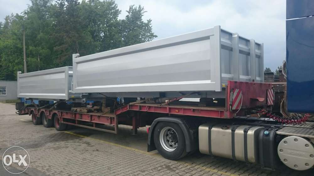 Ciężarówka z naczepą wywrotką w kolorze srebrnym, zaparkowana na placu. Widoczne koła, rama i fragment kabiny. Tło stanowią drzewa i budynek.