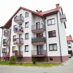 Nowoczesny, trzypiętrowy budynek mieszkalny z balkonami, wykończony w bieli i brązie, z widoczną anteną satelitarną na jednym z balkonów i suszącym się praniem na innym.