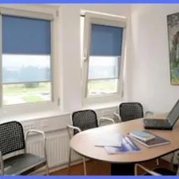 Wnętrze biura w Łodzi z dwoma oknami, w których zamontowano niebieskie rolety wewnętrzne. Na stole leży laptop i dokumenty, a pod oknami widać grzejnik.