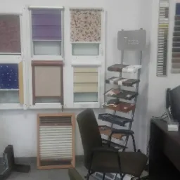 Ekspozycja różnych typów żaluzji i rolet okiennych w biurze, z próbkami materiałów i kolorów na ścianie, obok biurka z komputerem i regałem z dokumentami.