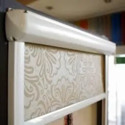 Zamknięta roleta plisowana w białej ramie, z beżowym materiałem w delikatny, roślinny wzór, zamontowana w oknie. Widoczny mechanizm rolety i fragment ściany w tle.