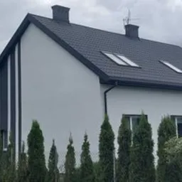 Nowoczesny dom jednorodzinny z jasną elewacją, ciemnym dachem i pionowym ciemnym pasem dekoracyjnym, otoczony tujami i zielenią, widok z zewnątrz w pochmurny dzień.