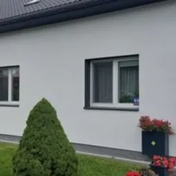 Biała, otynkowana ściana domu jednorodzinnego z trzema oknami o ciemnych ramach i balkonem z kwiatami, widok z poziomu trawnika z ozdobnymi krzewami i kwiatami w donicach.