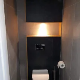 Nowoczesna toaleta z szarą płytką, podświetlaną wnęką z drewnianym obramowaniem, czarną ścianą z ukrytym systemem spłukiwania, oraz rolką papieru toaletowego i szczotką do WC