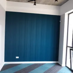 Ściana w kolorze głębokiego granatu z subtelną, pionową fakturą, biała listwa przypodłogowa, dywan w geometryczne pasy turkusu i szarości, surowy betonowy sufit z reflektorami, widok na okno.