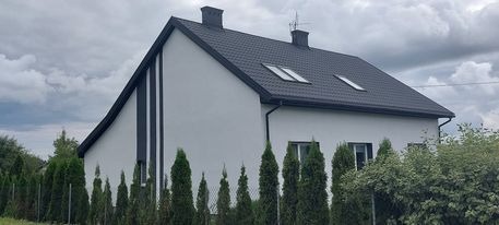 Nowoczesny dom jednorodzinny z jasną elewacją, ciemnym dachem i pionowym ciemnym pasem dekoracyjnym, otoczony tujami i zielenią, widok z zewnątrz w pochmurny dzień.