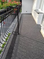 Balkon z szarą, kamienną posadzką, czarną, metalową balustradą i kwiatami w tle, widok z góry.