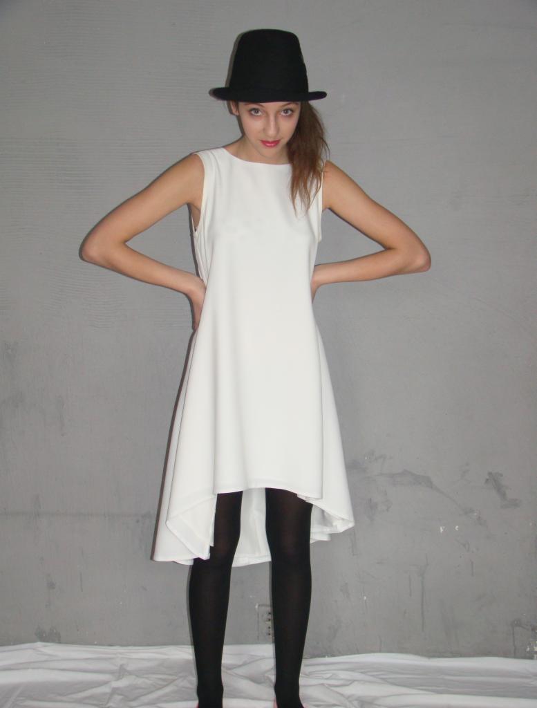 Młoda kobieta w białej, asymetrycznej sukience i czarnym kapeluszu pozuje na tle szarej ściany.